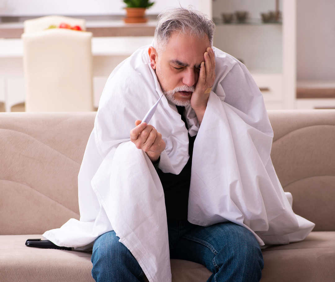 Symptome für Endokarditis: Mann sitzt mit Decke und Fieberthermometer auf dem Sofa.