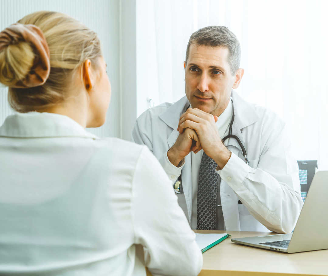 Ein Mann in weißem Kittel und eine Frau sitzen sich an einem Tisch gegenüber.