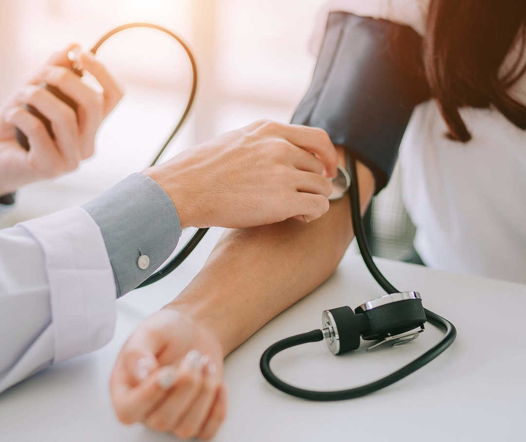Hypotonie-Betroffene sollten Ihren Blutdruck regelmäßig messen. Ärztin sitzt am Schreibtisch und misst den Blutdruck einer Patientin. Ist dieser zu niedrig, kann das auf eine Herzinsuffizienz hindeuten.
