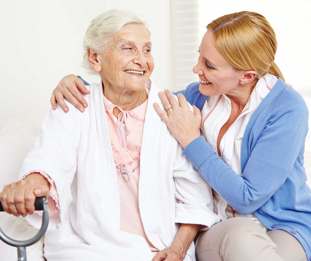 Ältere Herzinsuffizienz-Patientin sitzt neben ihrer Angehörigen, welche die Pflege übernimmt.