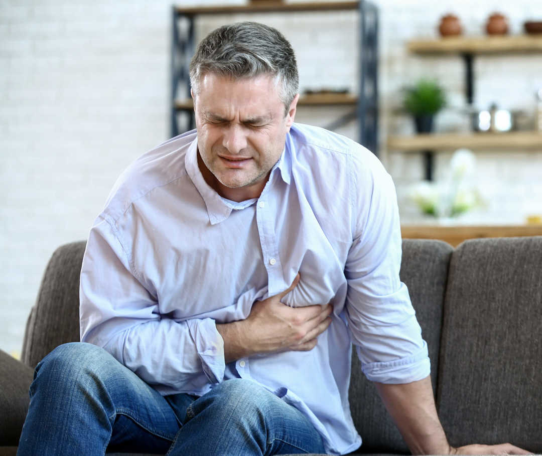 Mann sitzt auf dem Sofa und fasst aufgrund einer akuten Herzinsuffizienz an seine Brust.