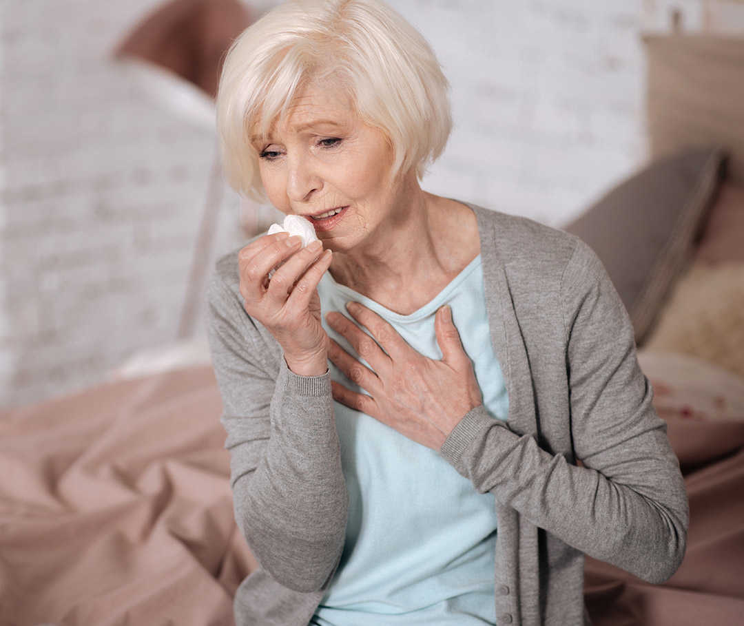 Frau hustet und hält sich die Hand auf die Brust, da sie durch COPD Probleme beim Atmen hat.