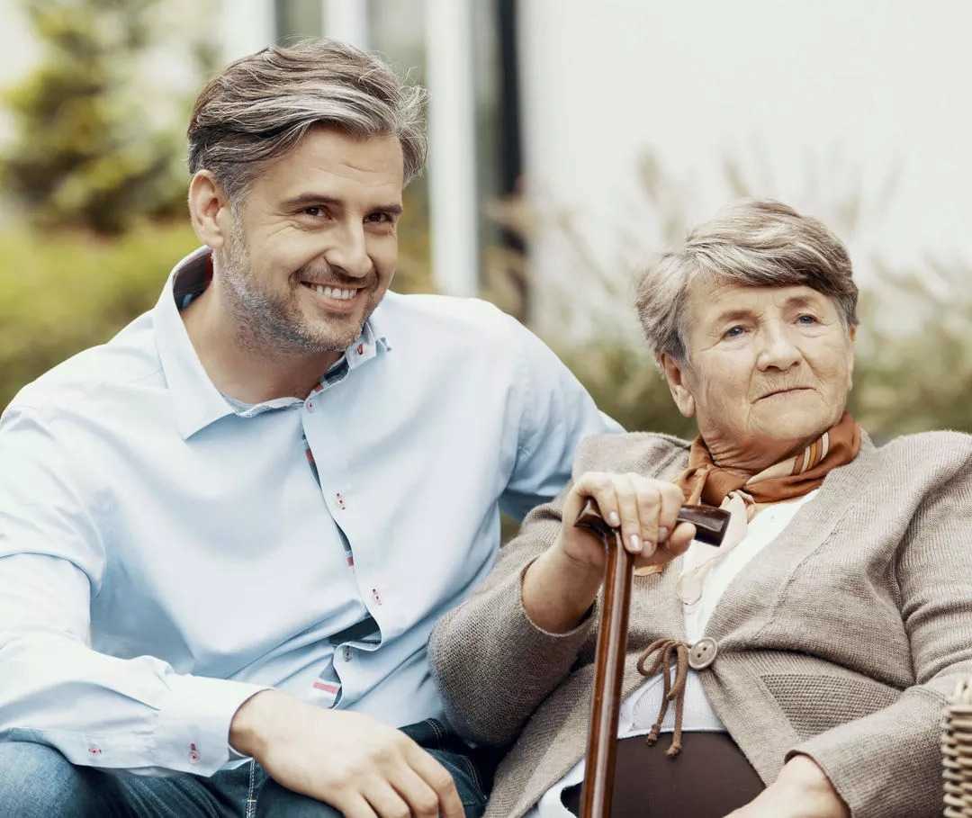 Sohn sitzt mit Mutter glücklich auf einer Bank im Garten, da sie die Anzeichen der vorliegenden Herzinsuffizienz rechtzeitig erkannt und in den Griff bekommen haben.