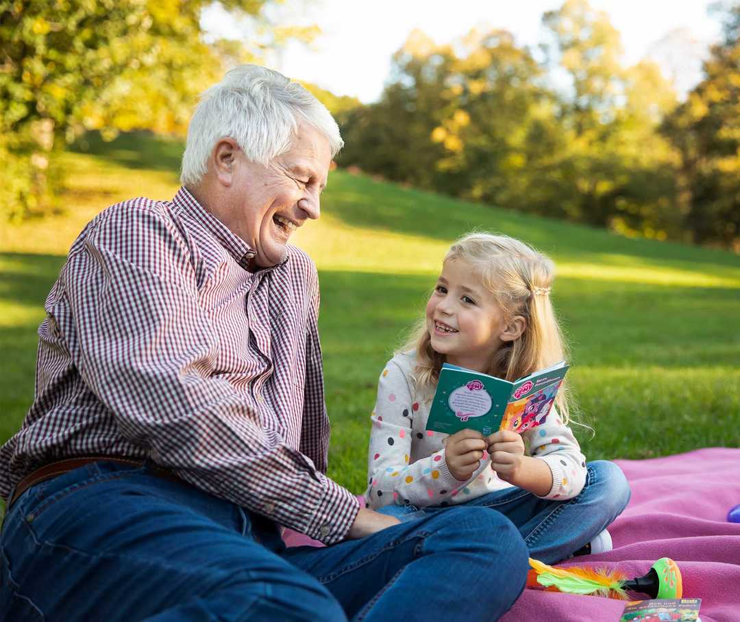 Älterer Mann sitzt mit Enkelin auf einer Decke im Grünen und vermeidet so Stress im Alltag.