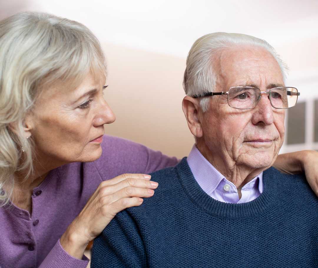 Eine ältere Frau beugt sich besorgt zu ihrem Mann, der in die Ferne schaut.