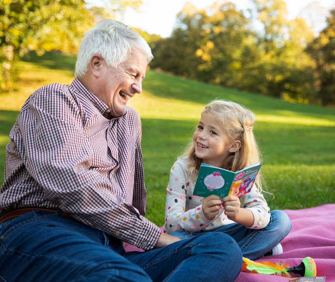 Ein älterer Mann sitzt lächelnd mit Enkelin auf einer Picknickdecke.