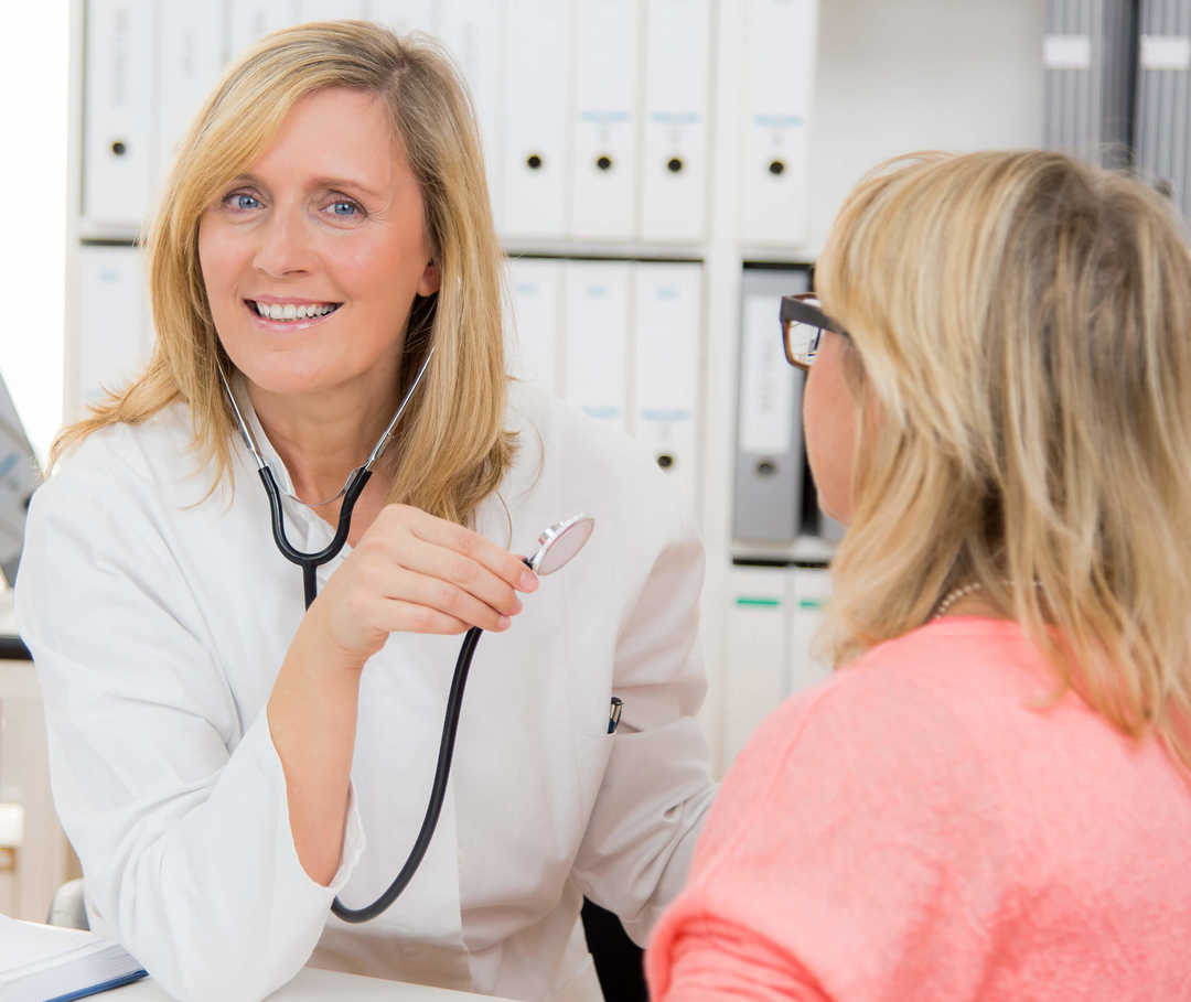 Regelmäßige Arztbesuche sind bei einer Herzinsuffizienz wichtig. Eine Frau in weißem Kittel sitzt einer weiteren Frau gegenüber und hat ein Stethoskop um den Hals und in der Hand.