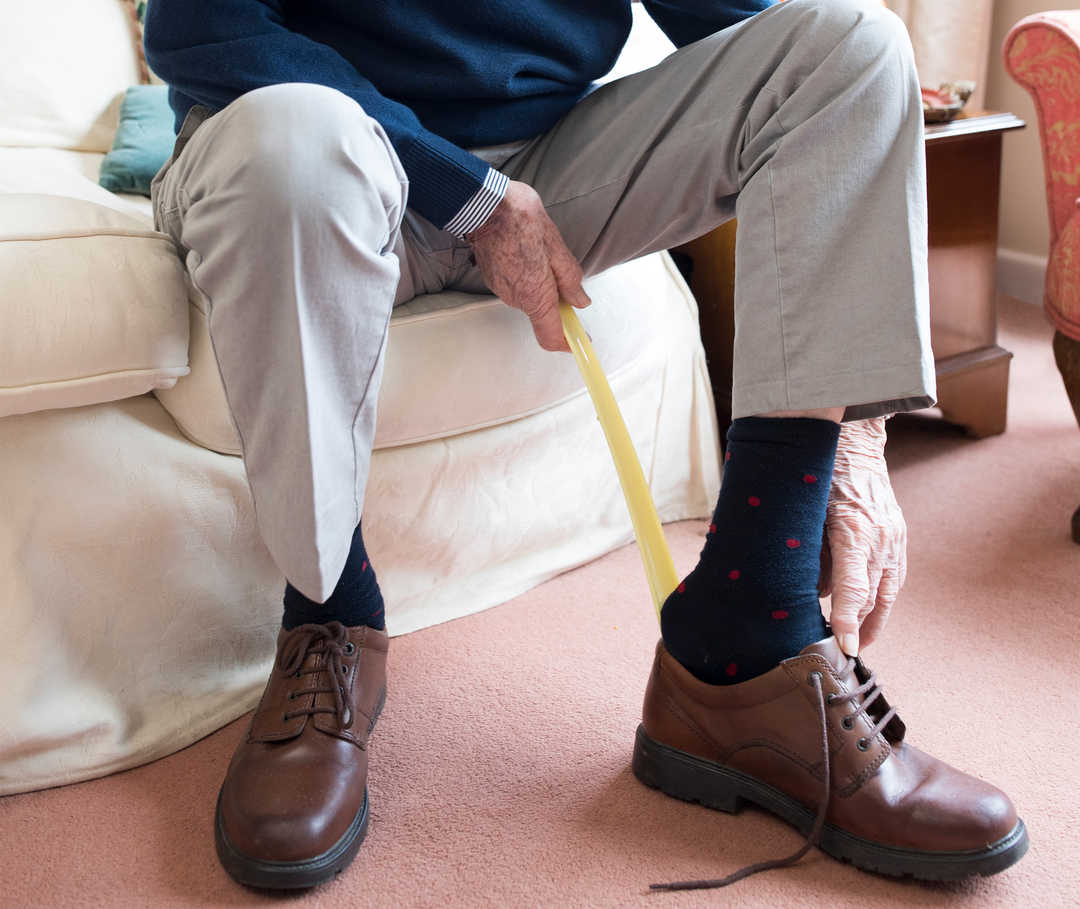 Mann zieht sich mit Schuhlöffel die Schuhe an und kommt nur schwer hinein, da seine Füße in der Folge einer Herzinsuffizienz angeschwollen sind.